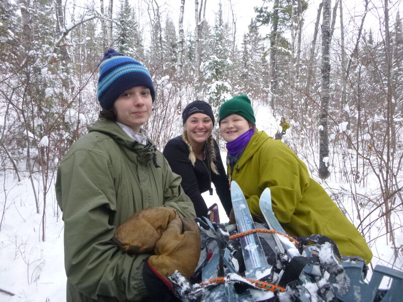 Three people are standing in a snowy forest. The person in the front is wearing a green jacket and a blue and black striped hat and holding a pair of snowshoes. Behind them to the left is a person wearing a black hat and a black jacket. To the right is a person wearing a green jacket and a green hat with a purple scarf. The background is a forest with snow-covered trees.